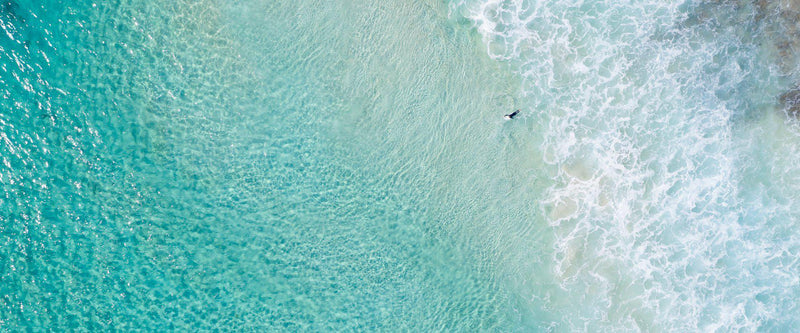 An aerial of a surfer paddling out through the clear waters of Western Australia.